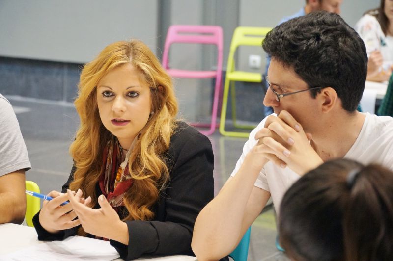 Una chica hablando a en una mesa varias personas sentadas a su lado Equipos